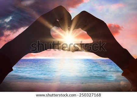 Woman making heart shape with hands against sunrise over magical sea