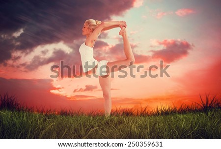 Sporty woman stretching body while balancing on one leg against red sky over grass