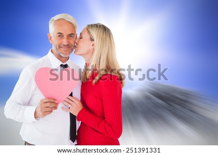 Handsome man holding paper heart getting a kiss from wife against large rock overlooking bright sky