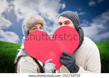 Attractive young couple in warm clothes holding red heart against green field under blue sky