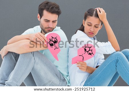 Sad couple sitting holding two halves of broken heart against grey
