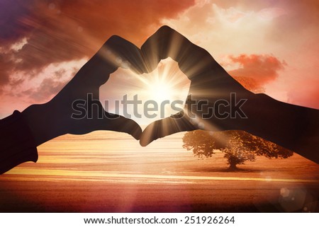 Couple making heart shape with hands against sunrise over field with tree