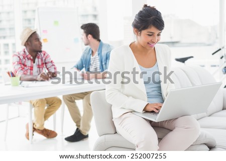 Smiling businesswoman using laptop on couch with colleagues behind her