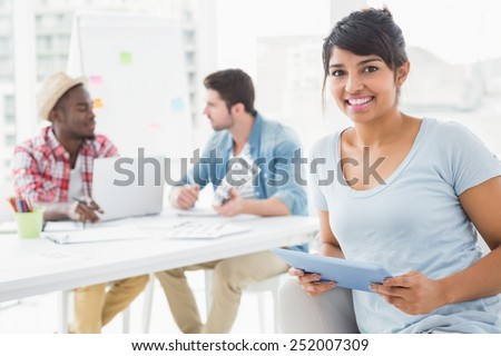 Smiling businesswoman holding tablet on couch with colleagues behind her