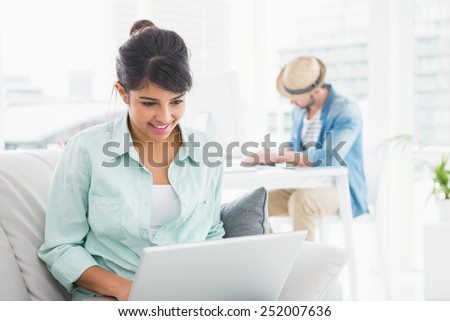Cheerful businesswoman using laptop on couch with colleague behind her