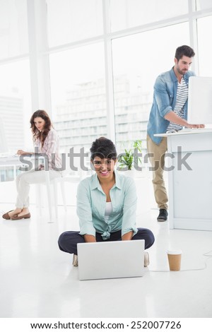 Happy businesswoman sitting on the floor using laptop with colleagues behind her
