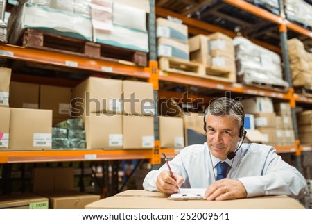Manager looking at camera while writing on clipboard in a large warehouse