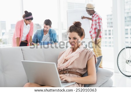 Happy businesswoman using laptop on couch with colleagues behind her