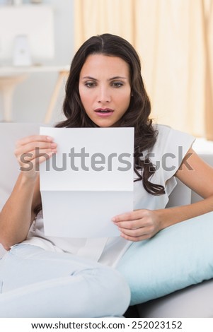 Shocked brunette reading letter on couch at home in the living room