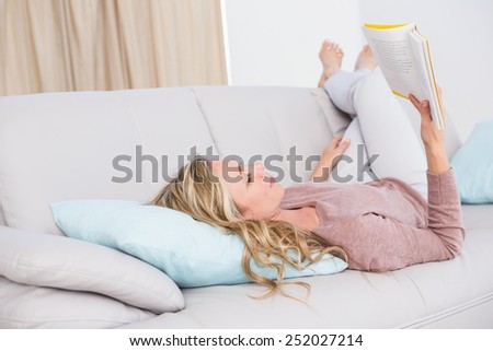 Petty blonde lying on couch reading book at home in the living room