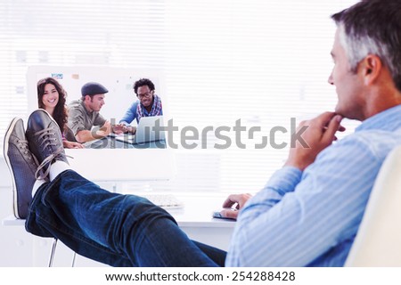 Relaxed man with feet on desk using computer against creative team working together with one smiling at camera