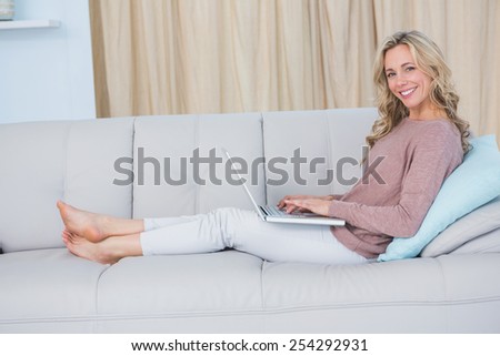Happy blonde on couch relaxing and using laptop at home in the living room