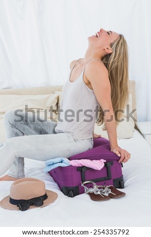 Smiling blonde closing baggage sitting on it in hotel room