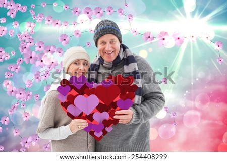 Happy mature couple in winter clothes holding red heart against digitally generated pretty flower background
