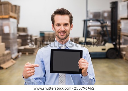 Warehouse manager showing tablet pc smiling at camera in a large warehouse