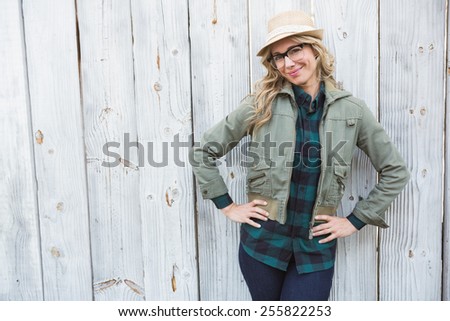 Smiling blonde in hat posing with hands on hips against bleached wooden planks