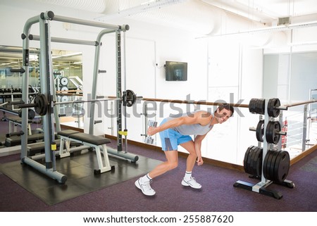 Smiling fit young man exercising with dumbbell against empty weights room with bench press