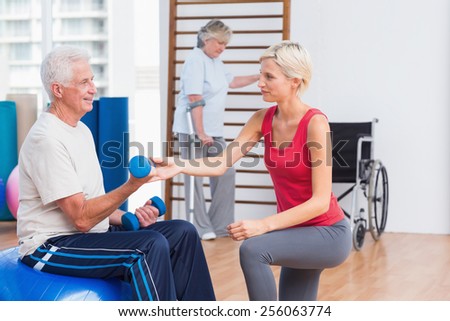 Female trainer assisting senior man in exercising with dumbbells while woman using crutches in background at gym