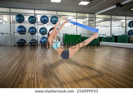 Fit young woman exercising with a blue yoga belt against large empty fitness studio with shelf of exercise balls