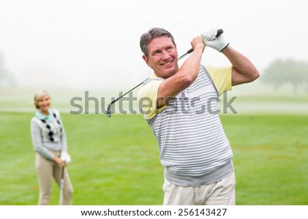 Happy golfer teeing off with partner behind him on a foggy day at the golf course