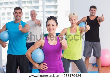 Portrait of happy female instructor with class holding medicine balls while gesturing thumbs up at gym