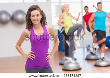Portrait of happy fit woman standing hands on hips with people exercising in background at gym
