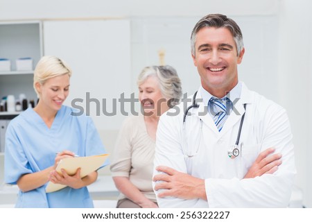 Portrait of confident doctor standing arms crossed while nurse and patient discussing in background at clinic