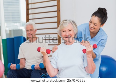 Portrait of happy senior woman lifting dumbbells while sitting with man and trainer in gym