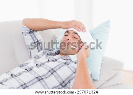 Sick man measuring temperature on thermometer while lying on sofa at home