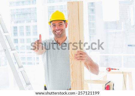Portrait of happy carpenter holding plank while gesturing thumbs up in building