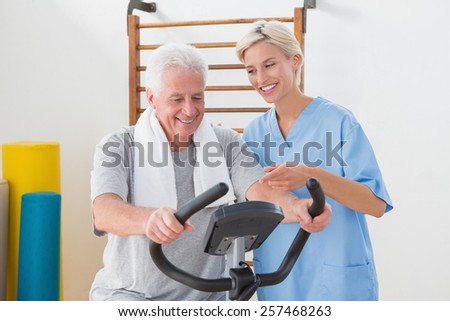 Senior man doing exercise bike with therapist in fitness studio