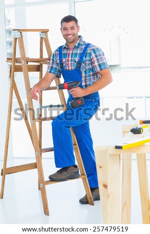Full length portrait of confident carpenter with power drill climbing ladder at construction site