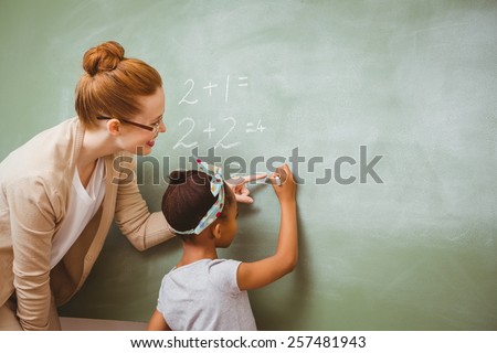 Rear view of teacher assisting little girl to write on blackboard in the classroom