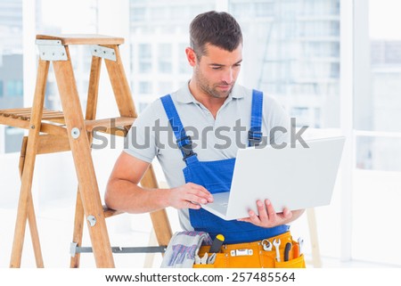 Repairman using laptop by step ladder in bright office