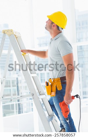 Side view of handyman with drill machine climbing ladder in building