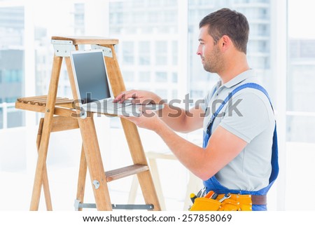 Side view of handyman using laptop by step ladder in bright office