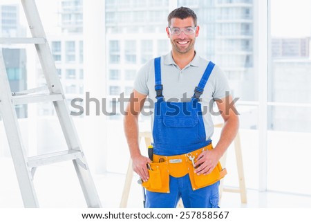 Portrait of smiling repairman in overalls standing hands on hips at bright office