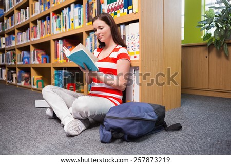 Pretty student sitting on floor reading book in library at the university