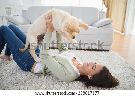 Happy young woman lifting puppy while lying on rug at home