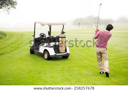 Golfer teeing off next to his golf buggy at the golf course