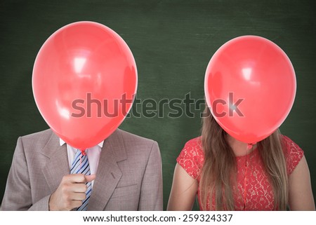 Geeky couple holding balloons in front of their faces against green chalkboard