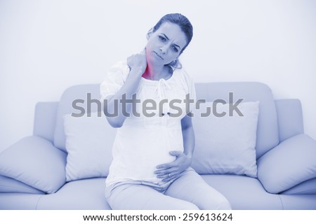 Attractive pregnant woman holding her injured neck while touching her belly and sitting on couch