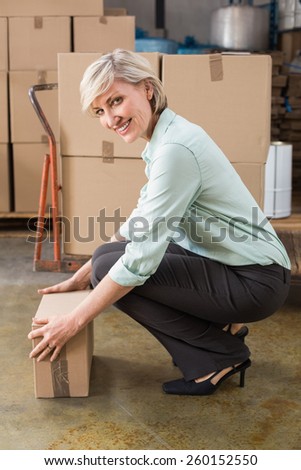 Smiling warehouse manager picking up cardboard box in a large warehouse