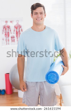 Happy young man holding exercise mat in medical office