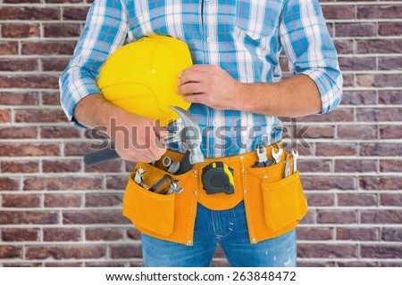 Manual worker wearing tool belt while holding hammer and helmet against red brick wall