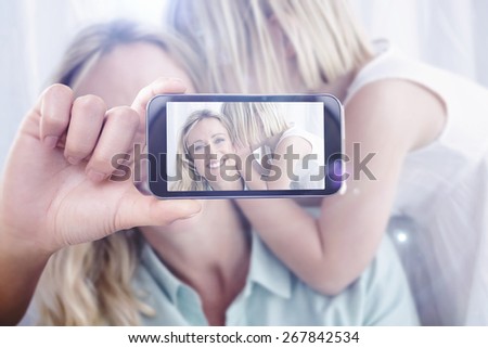 Hand holding smartphone showing against mother sitting with her daughter whispering a secret