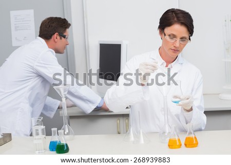 Scientist pouring chemical fluid in funnel and her colleague working with computer in laboratory