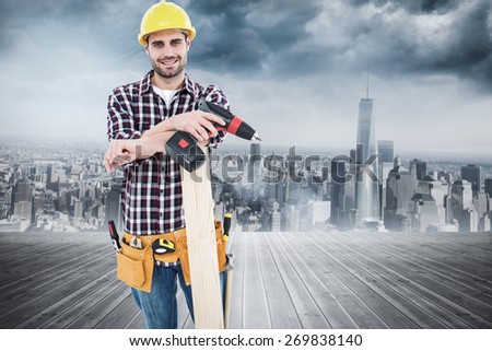 Confident male carpenter with drill machine and plank against room with large window looking on city