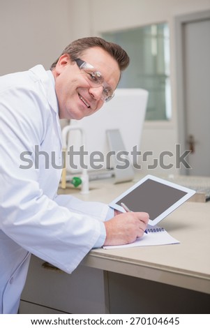 Smiling scientist holding tablet writing on notebook in the laboratory