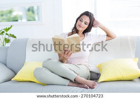 Smiling beautiful brunette relaxing and reading a book on the couch in the living room